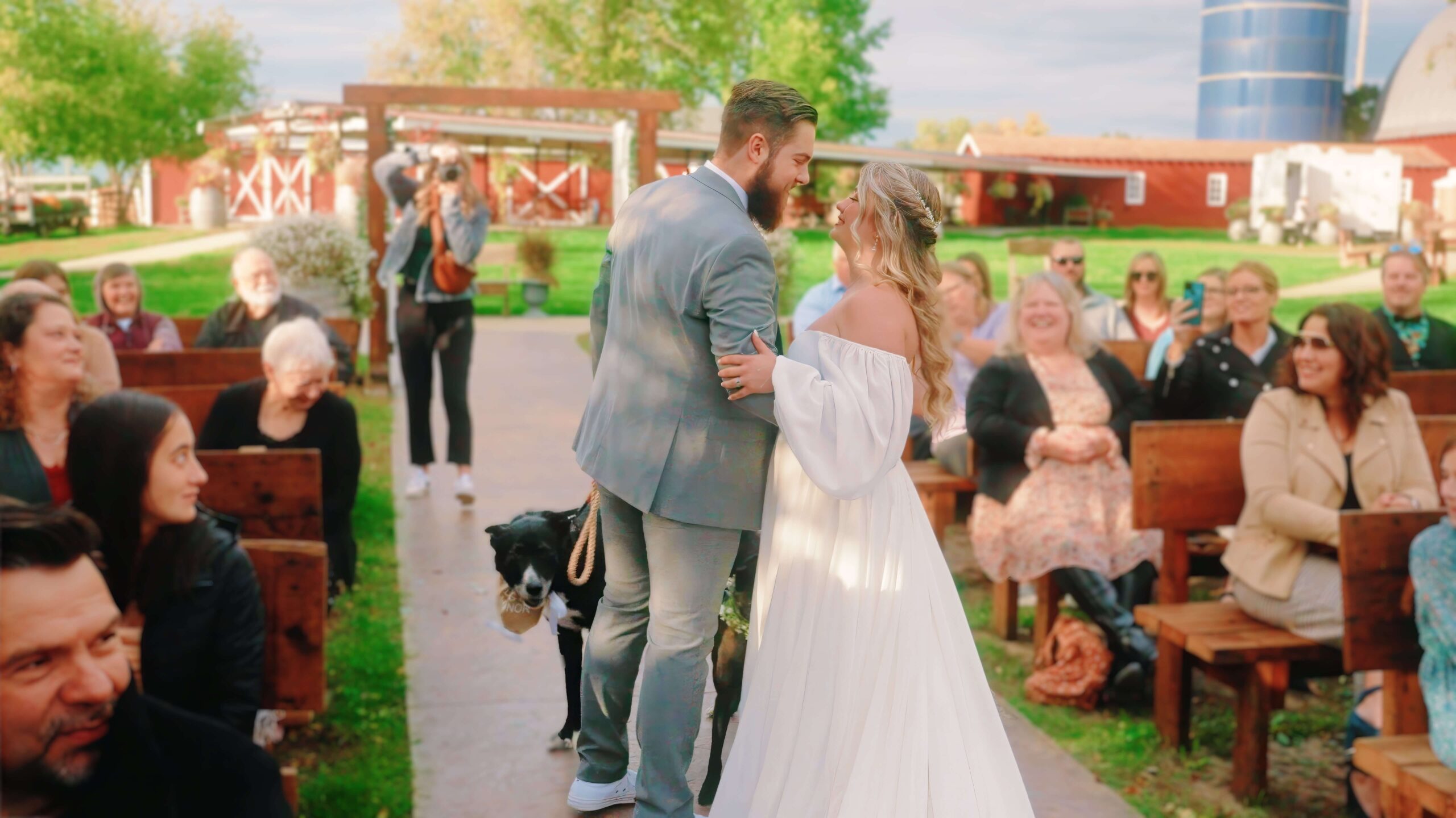 A couple walks arm in arm down an aisle outdoors, surrounded by seated guests. A dog on a leash walks beside them. The background includes green trees and red barns.