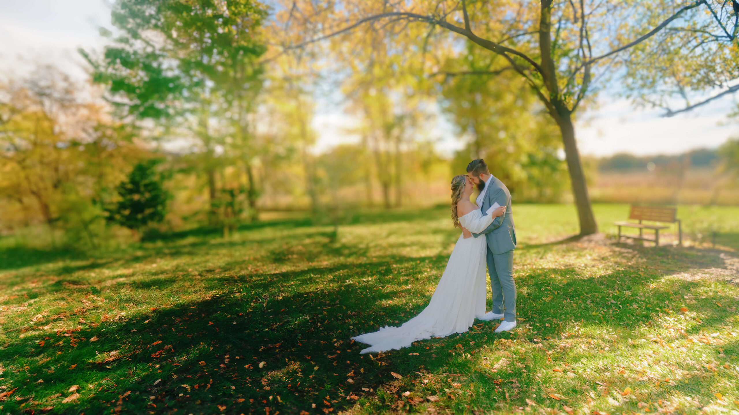 Bride and groom embrace in a lush green field with fall leaves on the ground and trees in the background during a fall wedding