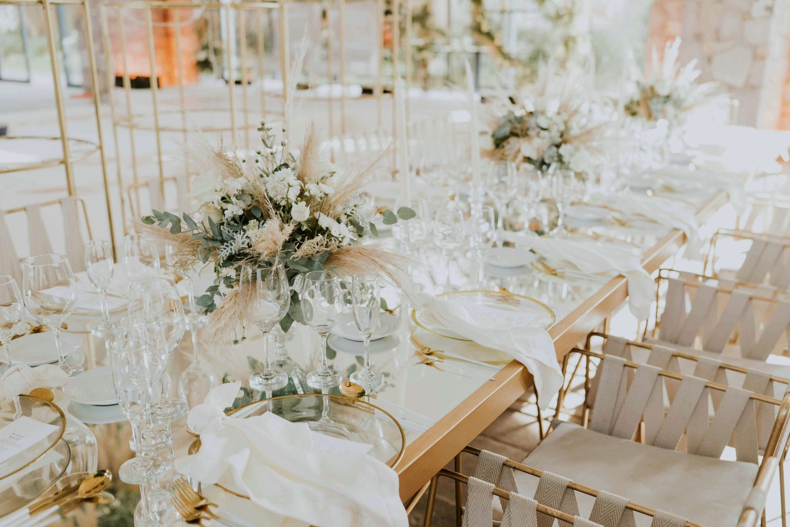 Elegant wedding reception table setting with pampas grass and white florals centerpiece, surrounded by glassware and gold accents.