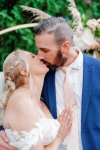 Bride and groom share a kiss under a wedding arch decorated with florals.