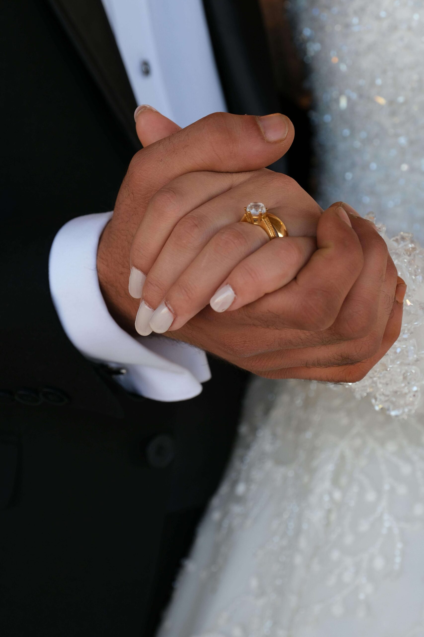Close-up of a couple holding hands at their Austin Country Club wedding, with one person wearing a sparkling white dress and an engagement ring with a gold band; both hands are gently clasped together.