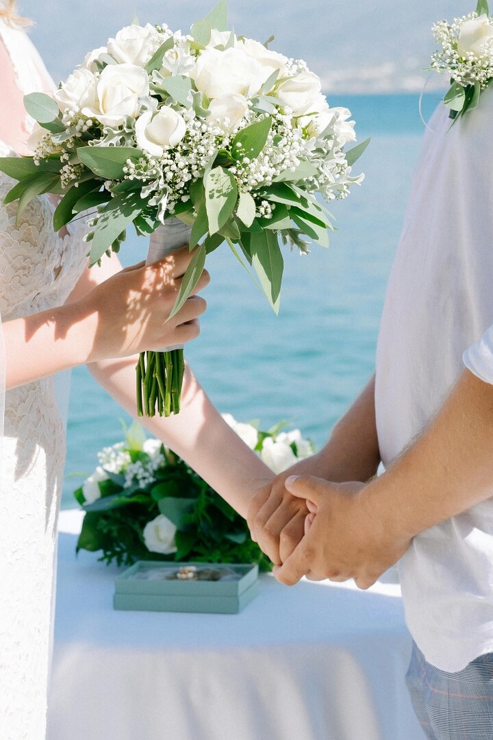 A bride holding a bouquet of white flowers and greenery stands with her groom, hand in hand near a table with wedding rings and blooms. The Grand Superior Lodge wedding scene features water and a stunning mountainous backdrop.