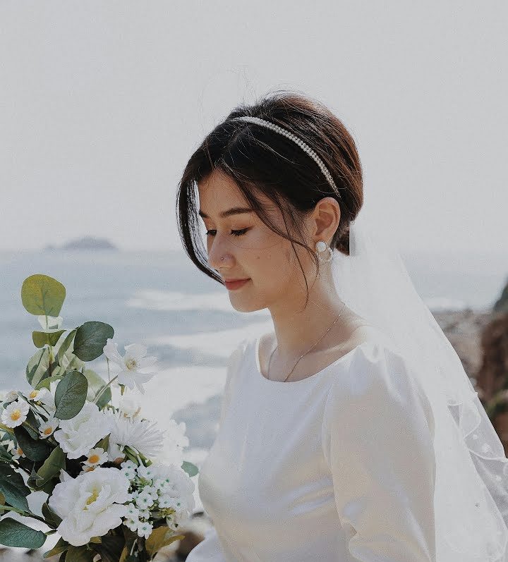 A bride in a long-sleeved white dress and veil stands outdoors at a Park Point Beach House wedding, holding a bouquet of white flowers and greenery, with the ocean and rocky shore visible in the background.